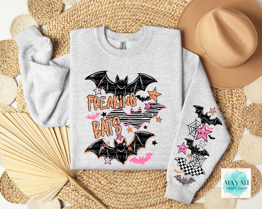 A light heather gray sweatshirt laid flat on a woven straw mat, featuring a Halloween-style bat graphic with the words “FREAKING BATS” in orange and pink, black bats, stars, stripes, and web motifs. A tan hat, folded straw fan, and circular woven decor surround the sweatshirt.