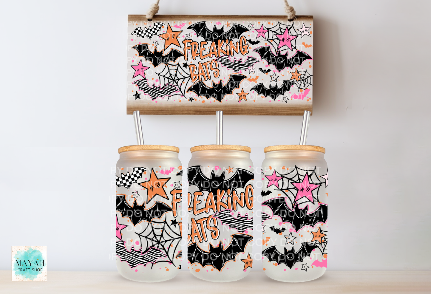 Three frosted glass mason jars with bamboo lids and metal straws, decorated with a black, white, pink, and orange 'Freaking Bats' Halloween pattern, displayed below a matching rectangular sign on a light wood surface against a white wall.
