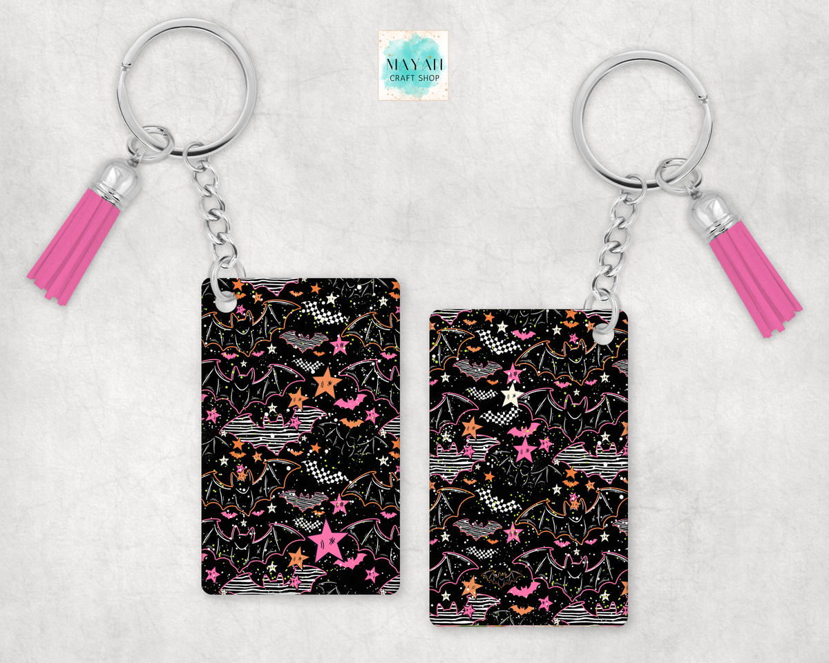 Two keychains with a black background and a colorful pattern of bats, stars, and sparkles, each attached to a silver key ring and a pink tassel charm, displayed on a light gray textured background.