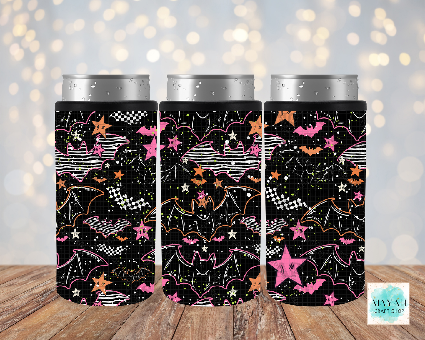 Three black can coolers with a Halloween-themed pattern of bats, stars, and checkered flags, shown on silver aluminum cans against a blurred bokeh background and wooden surface.