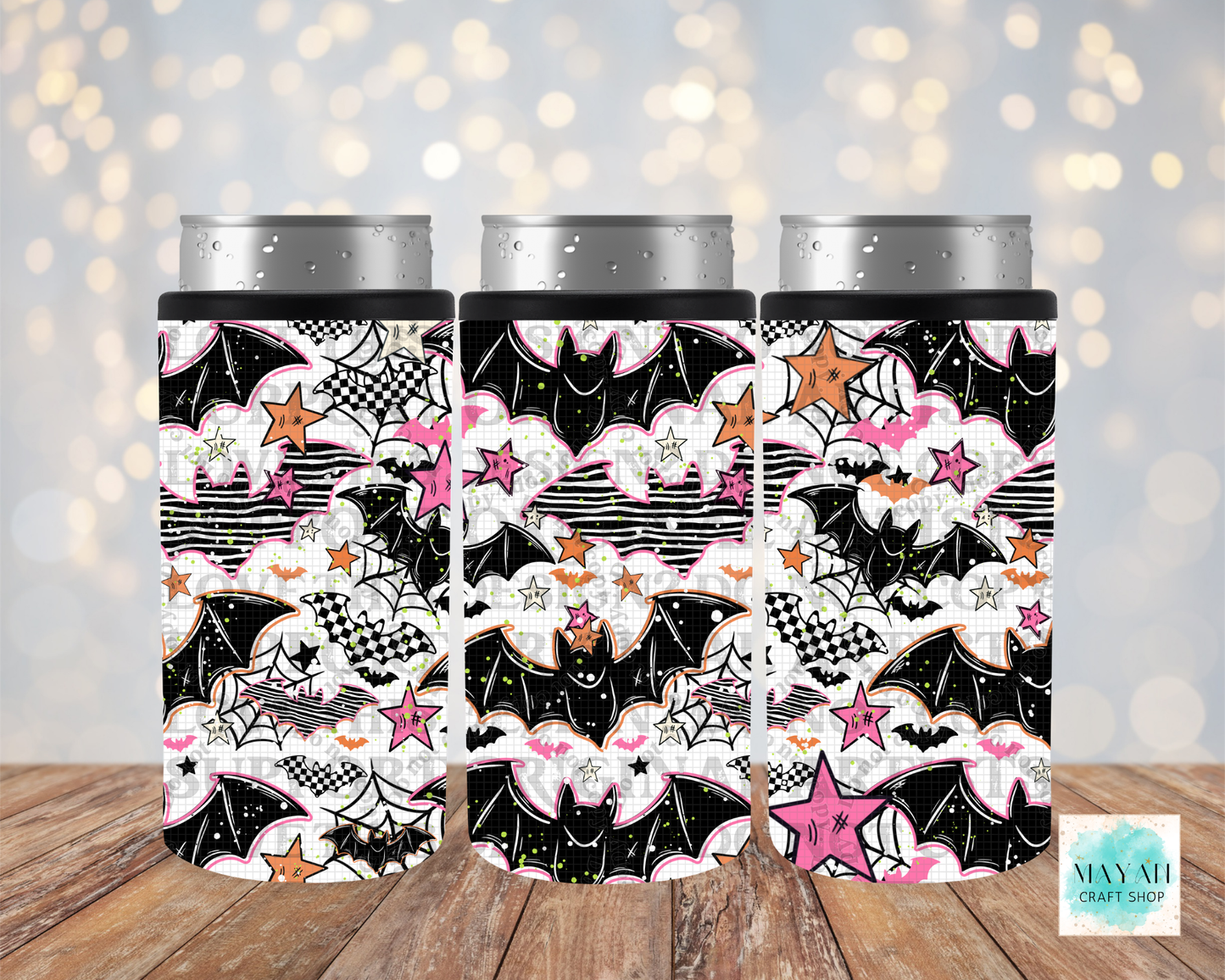Three slim can coolers with a Halloween-themed pattern featuring bats, spiders, webs, stars, and pink clouds, shown on silver aluminum cans against a blurred warm bokeh background and wood surface.