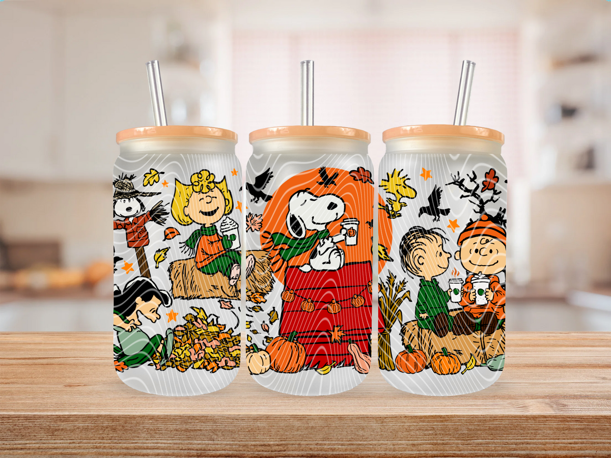 Three frosted glass cans with light wood lids and metal straws, printed with characters in an autumn Halloween scene, displayed on a wooden surface with a blurred kitchen background.