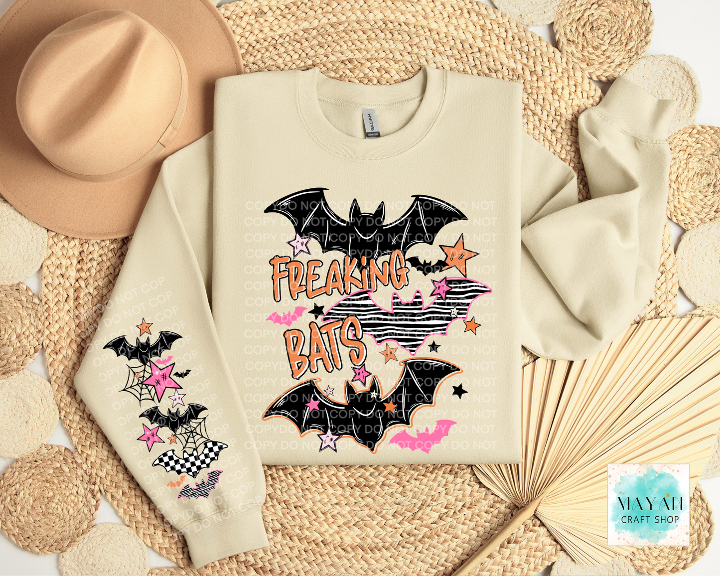 A cream-colored sweatshirt with a 'Freaking Bats' Halloween graphic, styled in a flat lay with a beige hat, woven placemats, and a paper fan. The graphic features black bats, pink stars, and orange text.
