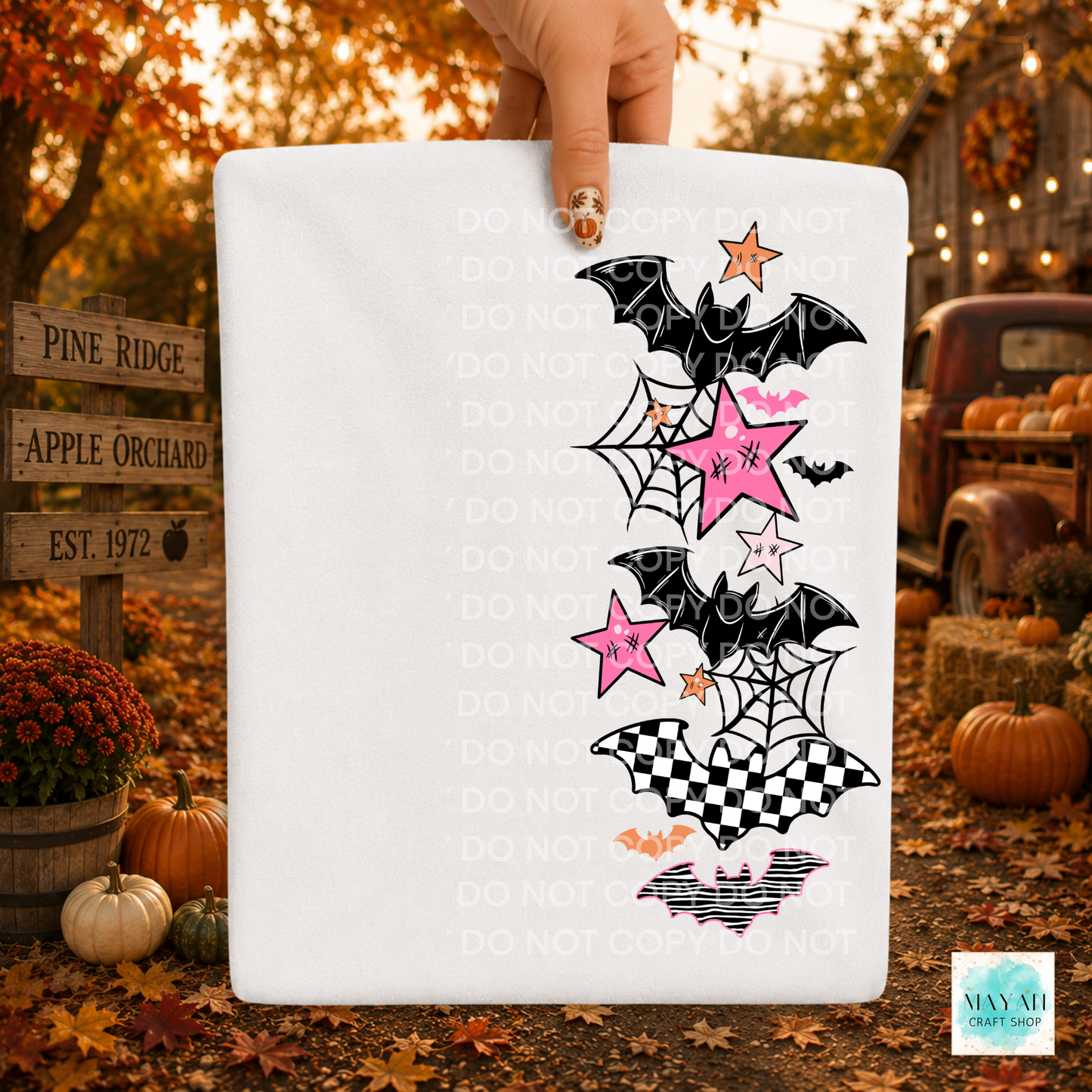 A white rectangular sign or board held by a hand, featuring a Halloween-themed graphic with black bats, pink and white stars, spiderwebs, and checkered shapes. The background is a fall yard scene with pumpkins, autumn leaves, and a rustic truck.