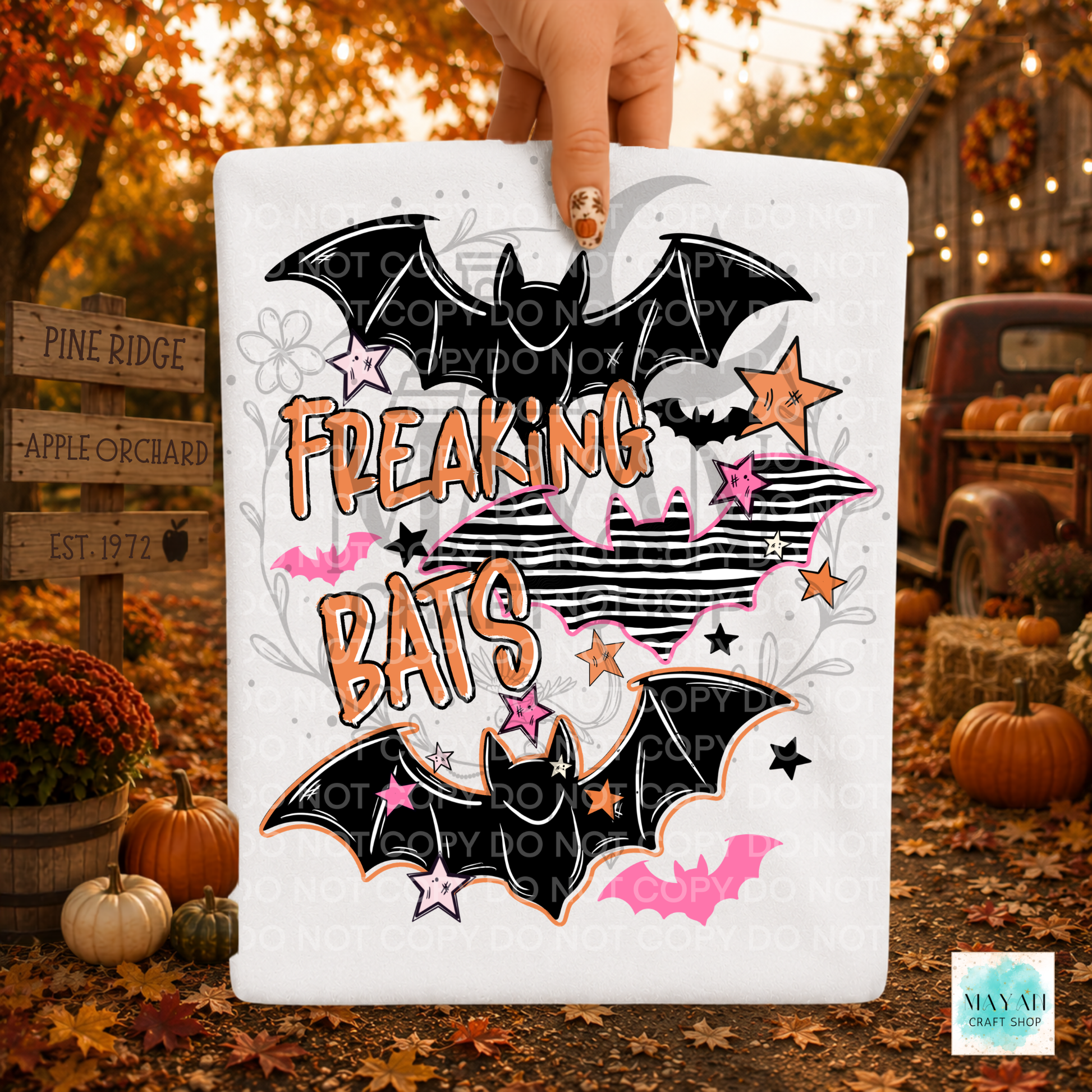 A white shirt held by a hand, featuring a Halloween-style graphic with two black bats, pink and orange text reading 'FREAKING BATS,' striped hearts, stars, and small bat silhouettes. The background is a fall yard scene with pumpkins, autumn leaves, a rustic truck, and wooden signs.