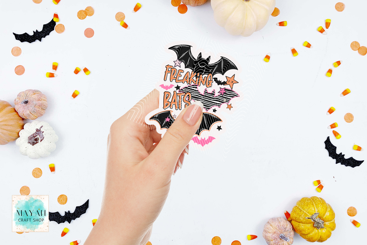 A hand holds a die-cut sticker featuring black bats, pink stars, and the text 'FREAKING BATS' in orange and pink. The background is a white flat-lay styled with small pumpkins, candy corn, and black bat cutouts.
