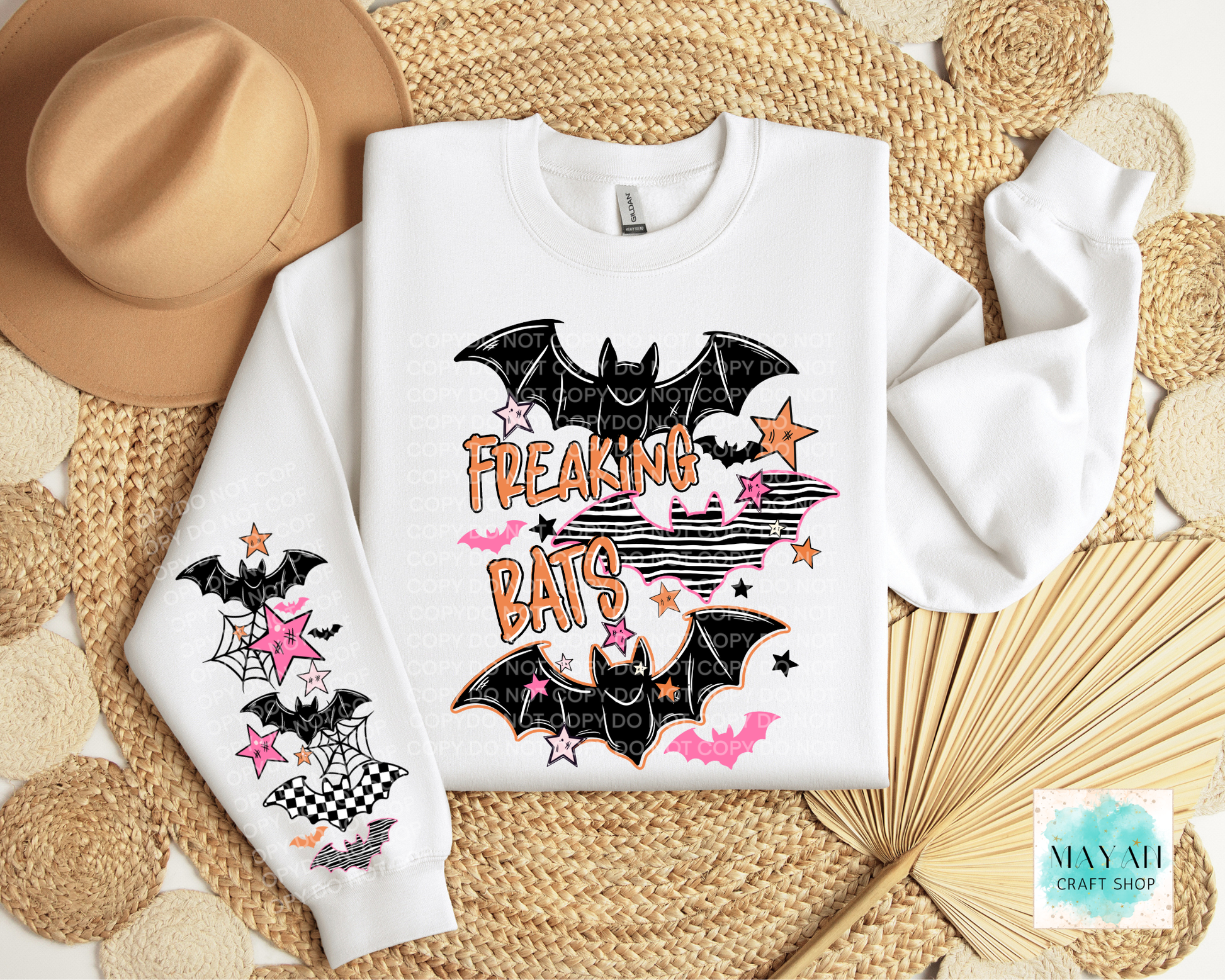 A white sweatshirt laid flat on a woven straw mat, decorated with black bat graphics, pink stars, and orange text reading 'FREAKING BATS.' A matching sleeve print is shown on the left, with a tan hat and dried palm leaves as styling props.