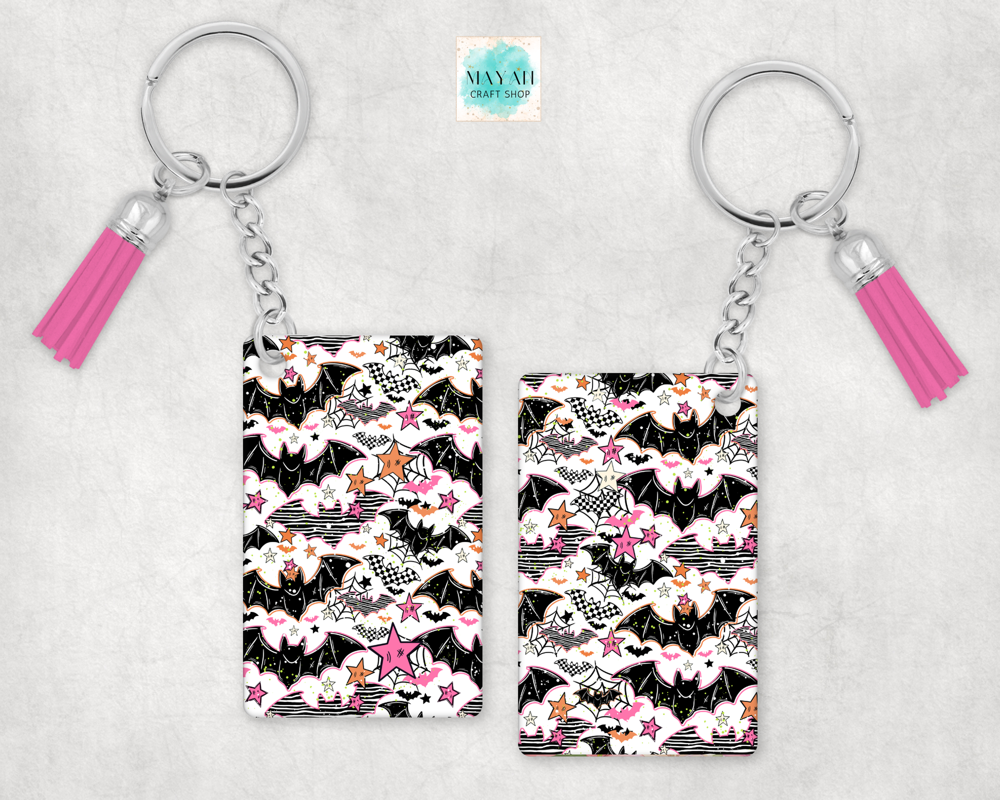 Two keychains with rectangular printed pendants and pink tassels, shown on a light gray textured background. The pendants feature a black, white, and pink pattern with bats, stars, and clouds.