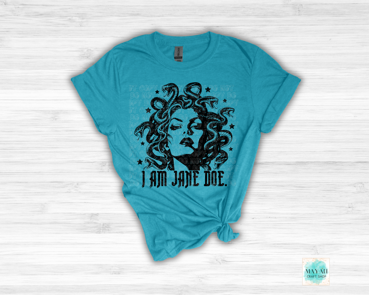 A heather galapagos graphic t-shirt featuring a black illustration of Medusa with snakes and the text 'I AM JANE DOE.' printed on the front, styled flat on a white wood background.