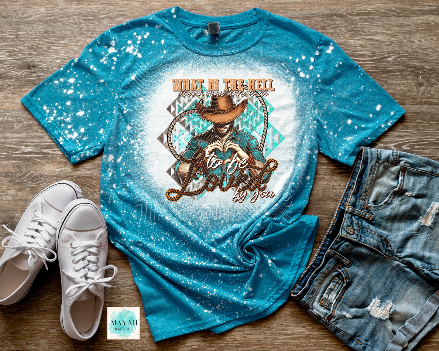 Blue graphic t-shirt with cowboy design and text on a wooden surface with white sneakers and denim shorts.