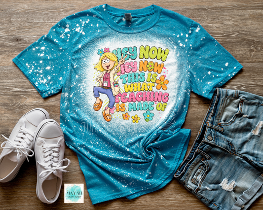 A turquoise blue tie-dye graphic t-shirt with a cartoon girl and the text 'HEY NOW I'S NOW THIS IS WHAT TEACHING IS MADE OF', styled in a flat lay on a wooden surface with white sneakers, distressed denim shorts.