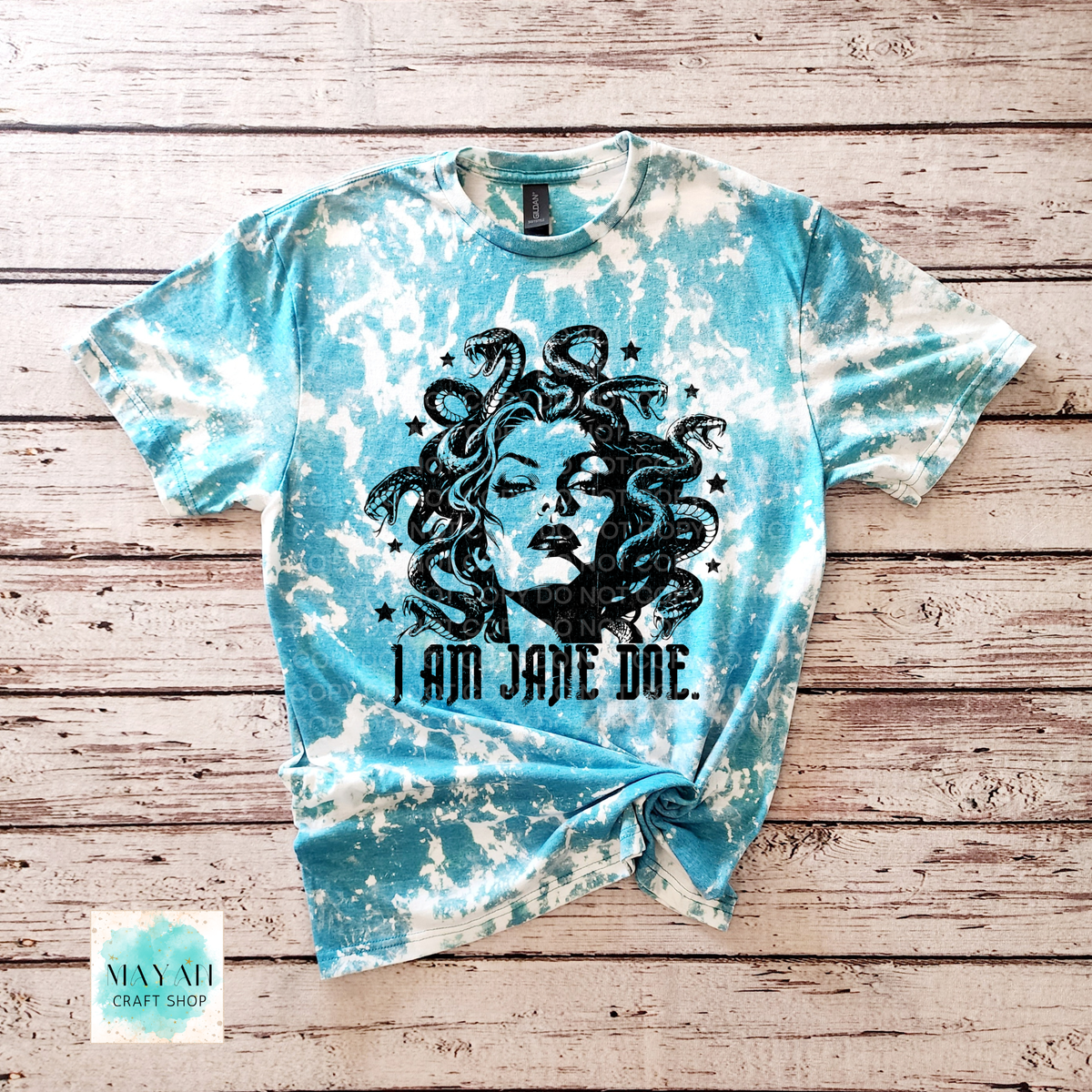 A short-sleeve t-shirt, heather galapagos bleached, featuring a black graphic of Medusa's hair and the text 'I AM JANE DOE.' It is laid flat on a rustic wooden plank background.