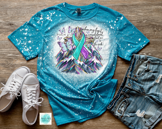 A flat-lay of a teal short-sleeve t-shirt with a colorful mountain and ribbon graphic, styled with white sneakers and distressed denim shorts on a wood background.