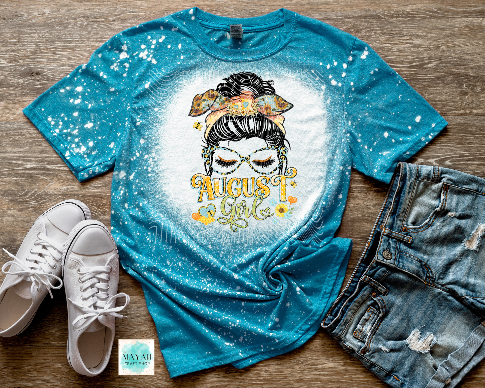 A turquoise blue short-sleeve graphic T-shirt laid flat on a wood surface, styled with white sneakers, distressed denim shorts, and a small card. The shirt has a white distressed center and a graphic of a woman with a headband and the words 'August 1st' in yellow/orange.
