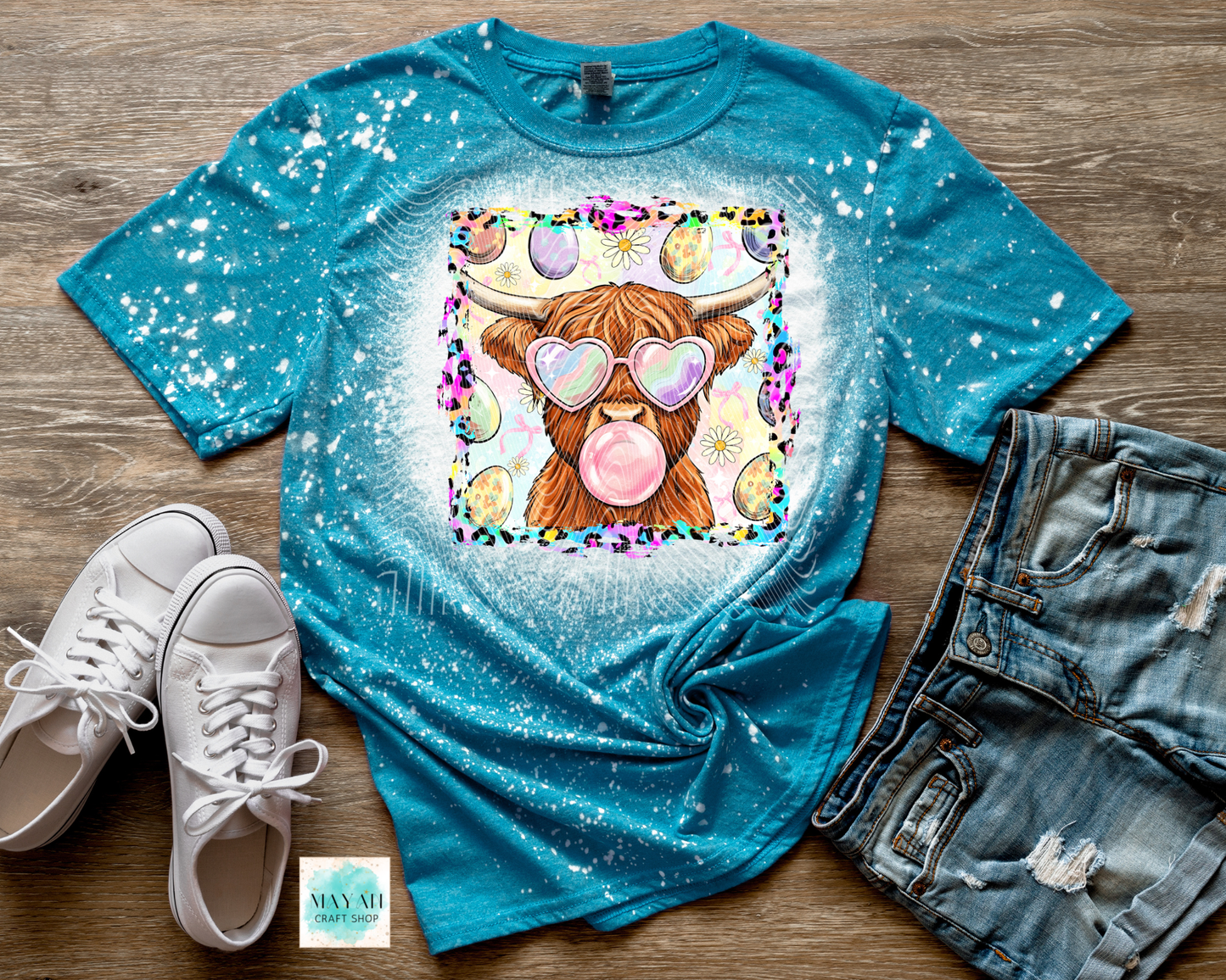 Blue t-shirt with a cartoon cow blowing a bubble, paired with white sneakers and denim shorts on a wooden surface.