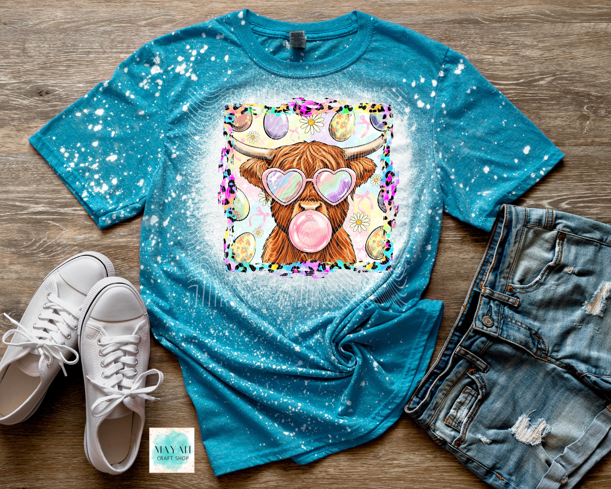 Blue t-shirt with a cartoon cow blowing a bubble, paired with white sneakers and denim shorts on a wooden surface.