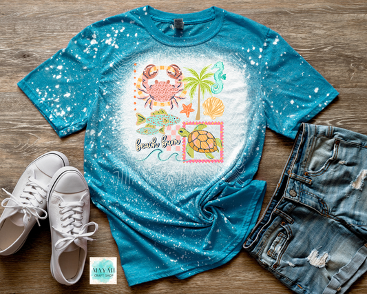 Blue t-shirt with beach-themed design on a wooden surface with white sneakers and denim shorts.