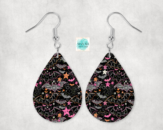 A pair of teardrop-shaped dangle earrings with a black background and a colorful pattern of pink stars, orange stars, white stars, and whimsical doodles. The earrings are suspended from silver fishhook wires.