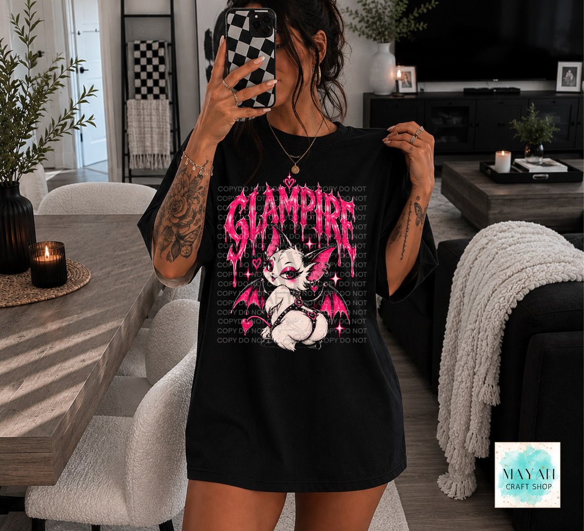 A woman takes a mirror selfie in a modern living room while wearing an oversized black graphic T-shirt. The shirt features a pink and white cartoon vampire design with the word 'GLAMPERE' in dripping pink letters.