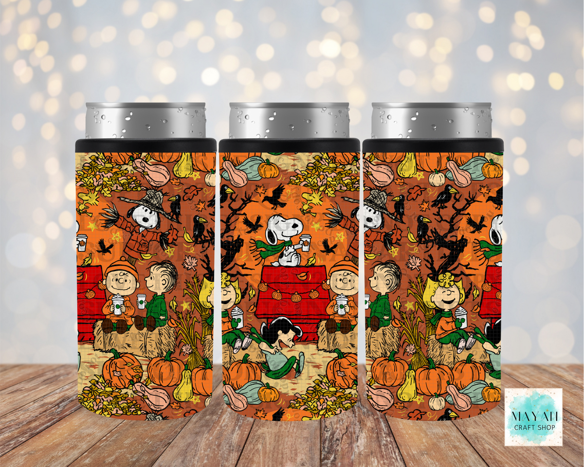 Three insulated can coolers with silver metal can tops, featuring a colorful autumn-themed Peanuts print with pumpkins and characters on a wooden surface against a blurred bokeh background.