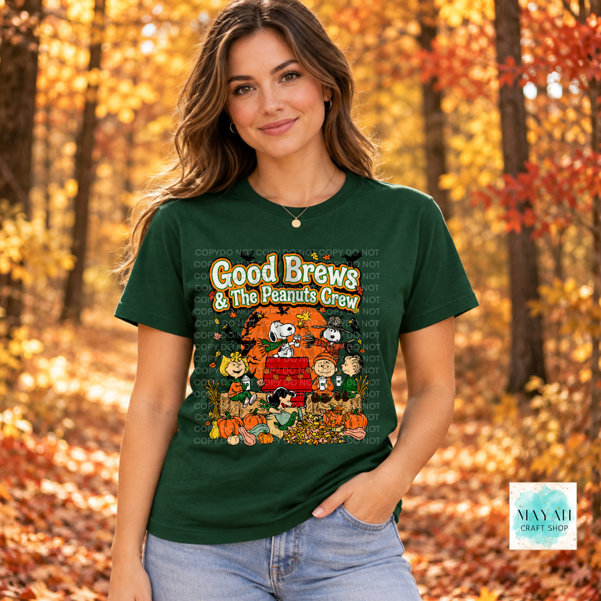A woman wearing a dark green graphic t-shirt featuring the 'Good Brews' design with vintage characters, set against an autumn forest background.