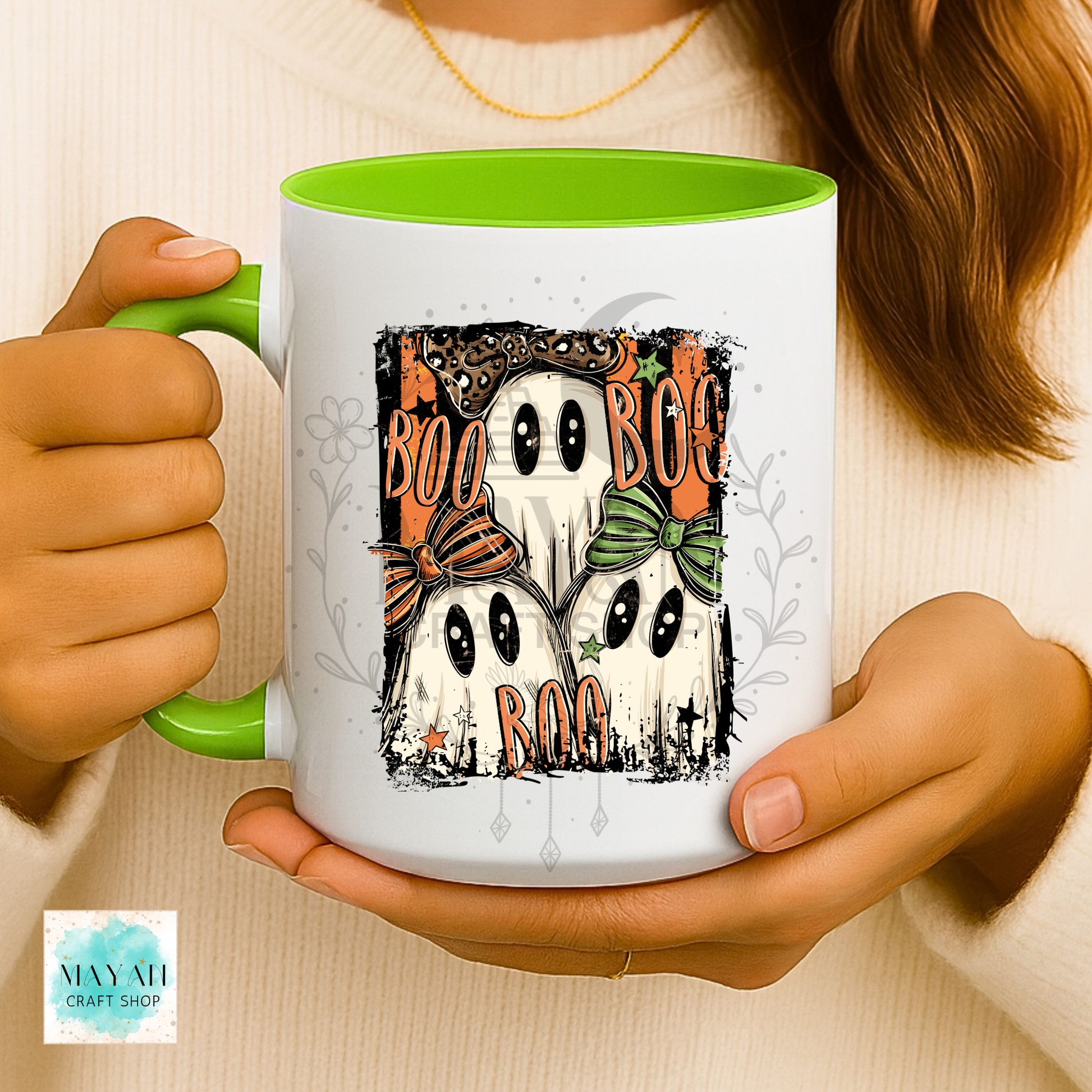 A person holds a white ceramic mug with a bright green handle and interior. The mug features a Halloween graphic with three cartoon ghosts, orange 'BOO' text, and gray decorative details.