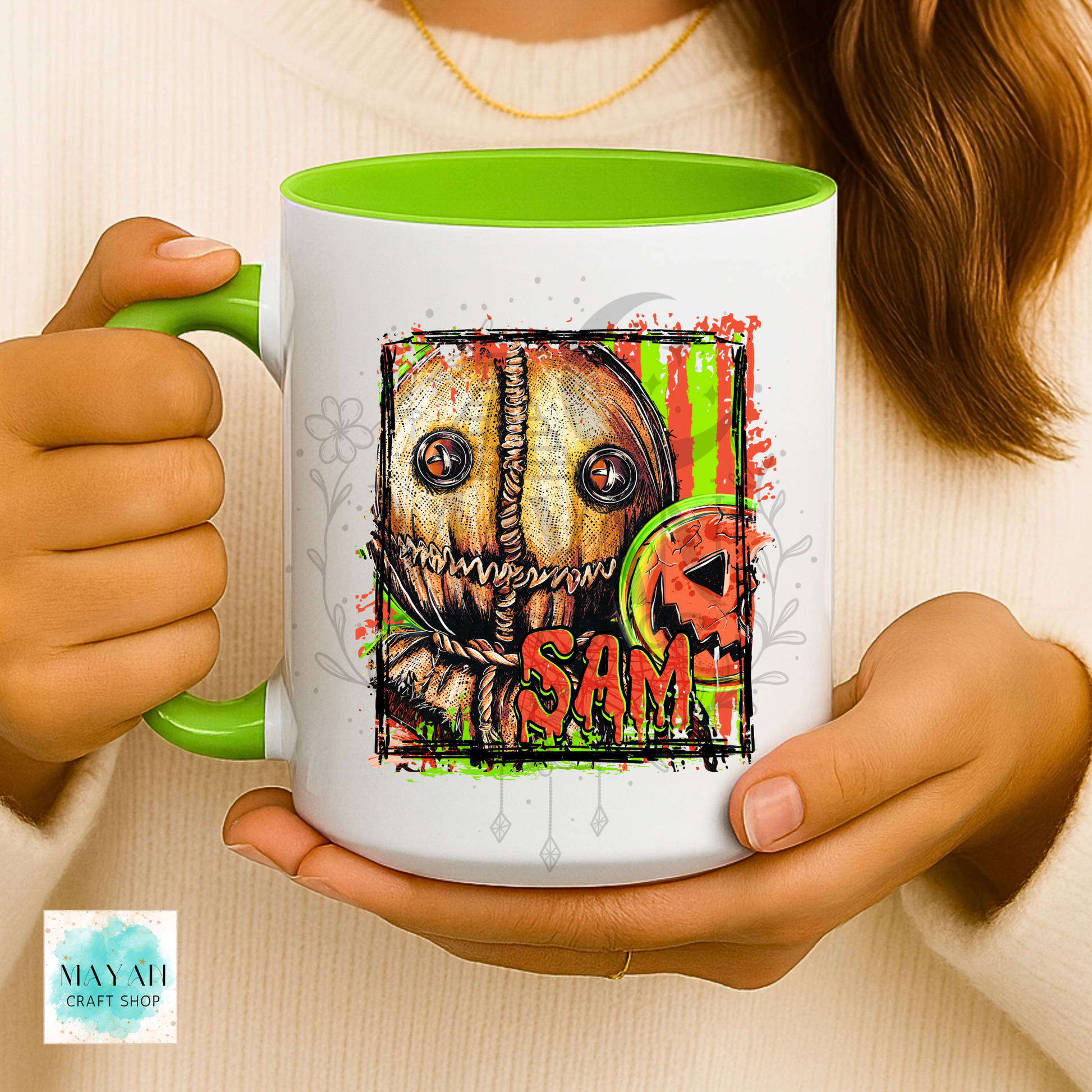 A person holds a white ceramic mug with a bright green handle and interior. The mug features a colorful graphic of a stitched horror character with a name.