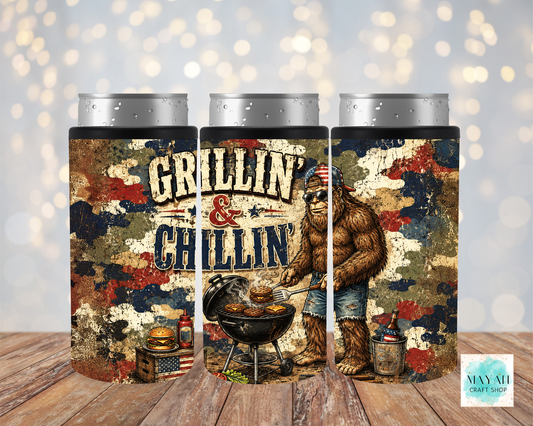 Three cylindrical can cooler with a creature grilling design and 'Grillin' & Chillin'' text on a wooden surface.