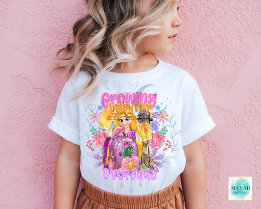 A child wearing a white graphic T-shirt featuring a princess with the text 'Growing brighter' in pink and yellow, posed against a pink textured wall.
