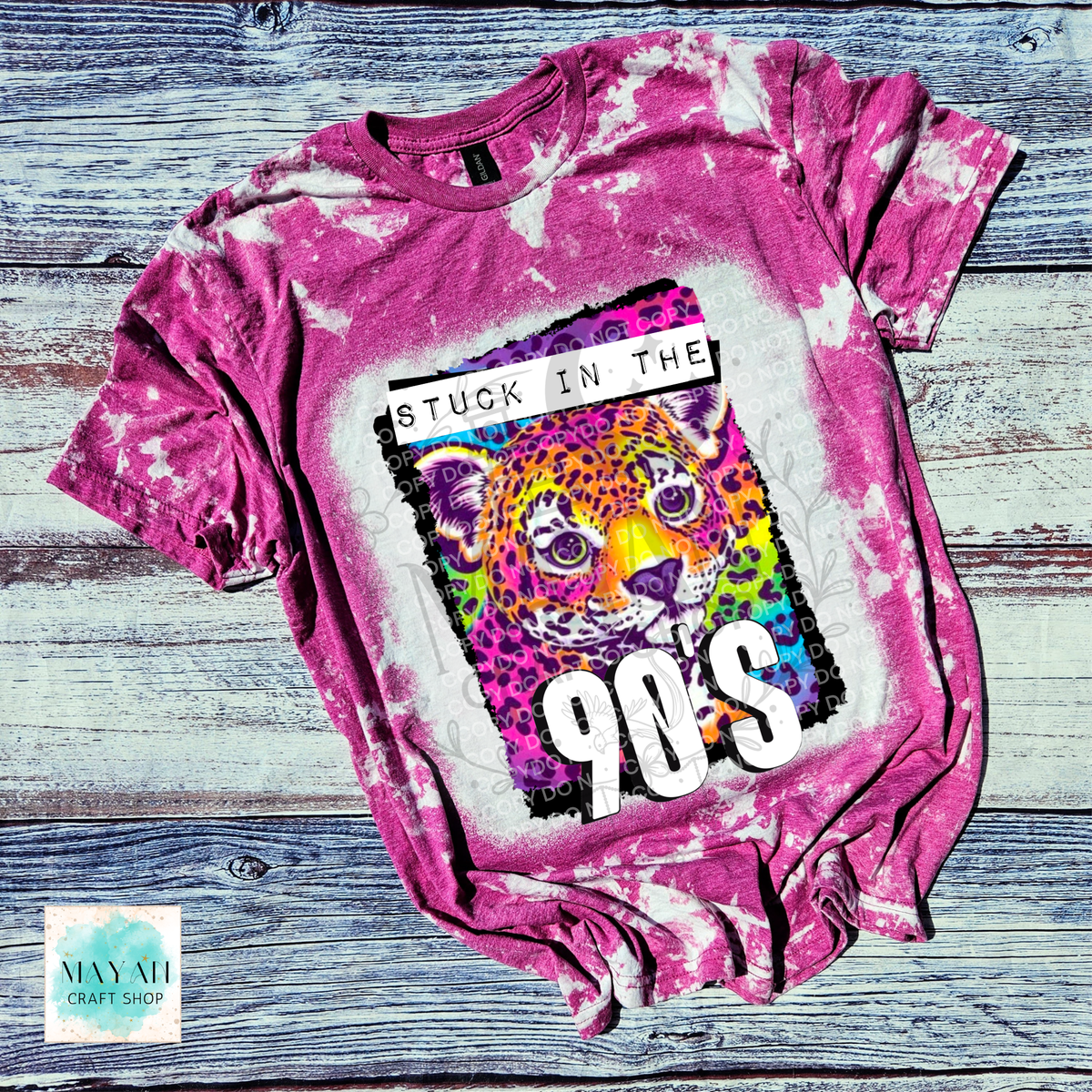 A berry bleached graphic t-shirt laid flat on a rustic blue-gray wooden plank background. The shirt features a central graphic of a colorful leopard with the text 'STUCK IN THE 90s'.