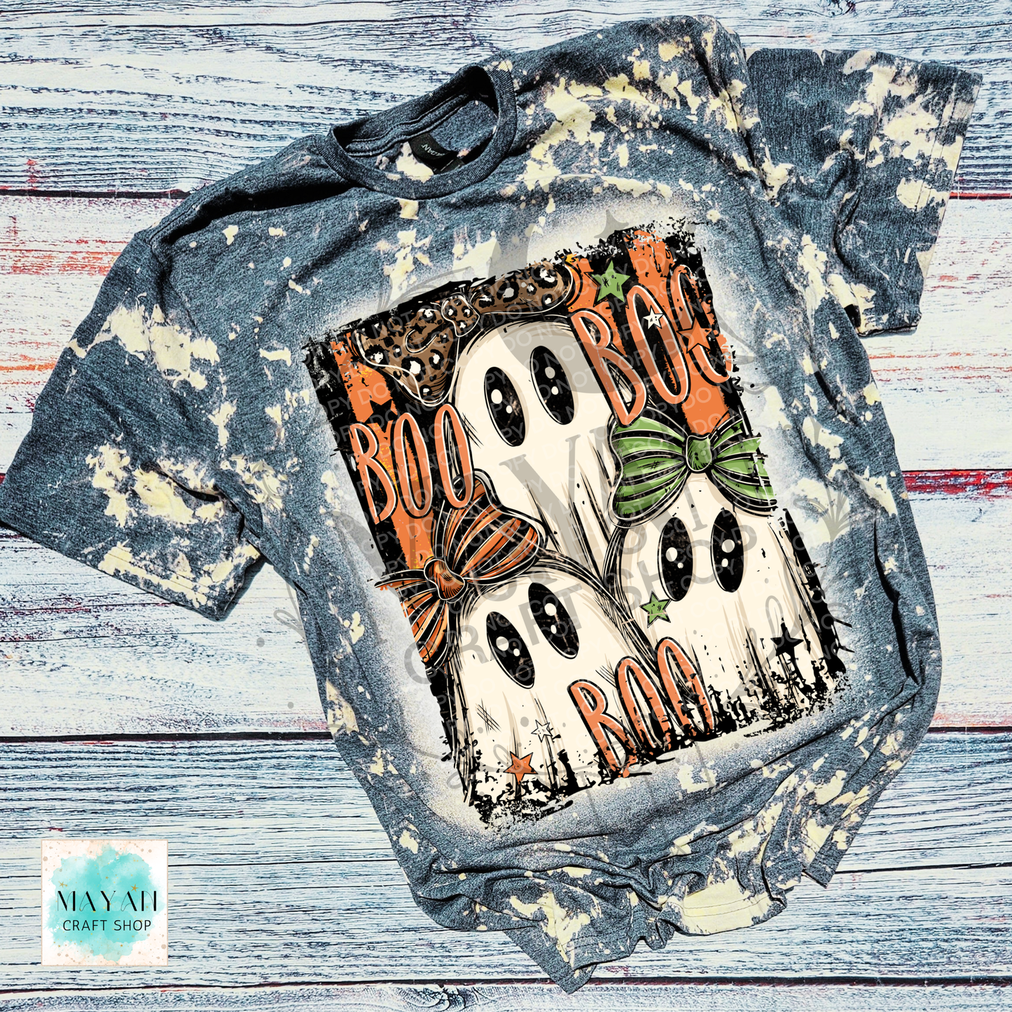 A short-sleeve graphic T-shirt laid flat on a distressed whitewashed wood background. The shirt has a blue-gray acid wash with cream splatter effects and features a large Halloween ghost graphic on the front with the words “BOO BOG BOO” in orange, plus orange and green bows, stars, and black outlines.