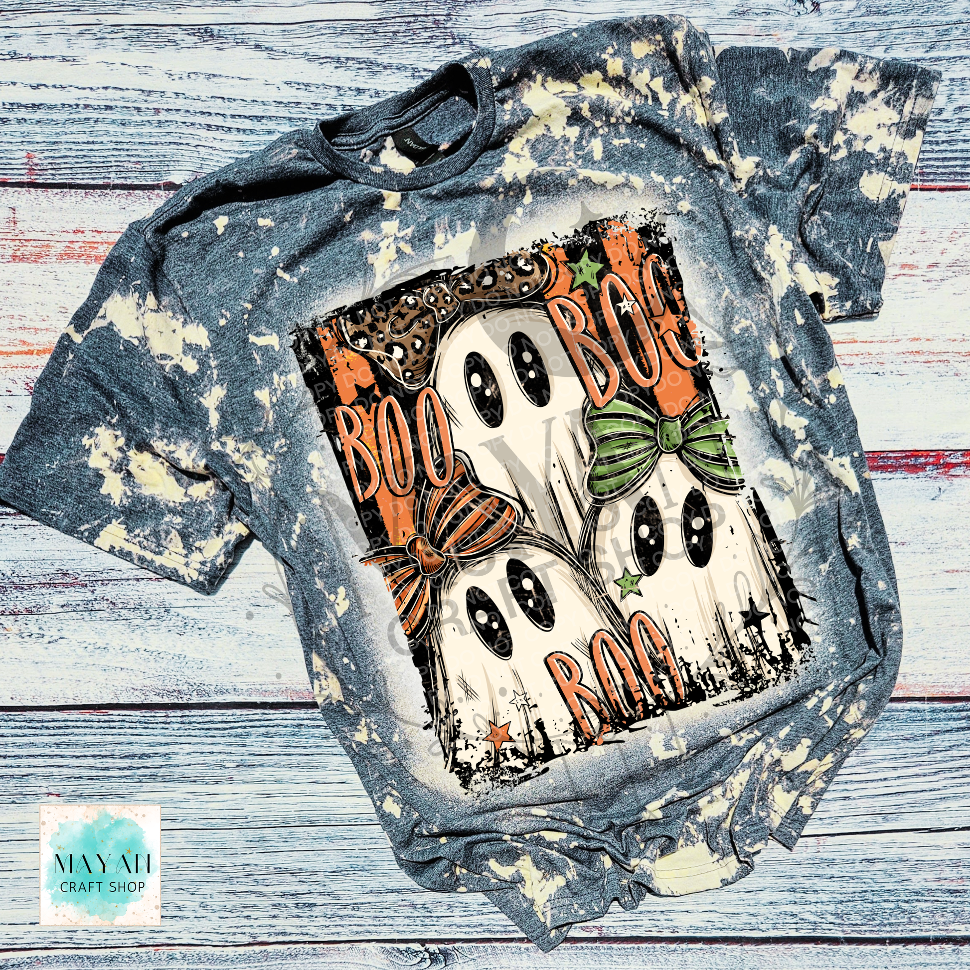 A short-sleeve graphic T-shirt laid flat on a distressed whitewashed wood background. The shirt has a blue-gray acid wash with cream splatter effects and features a large Halloween ghost graphic on the front with the words “BOO BOG BOO” in orange, plus orange and green bows, stars, and black outlines.