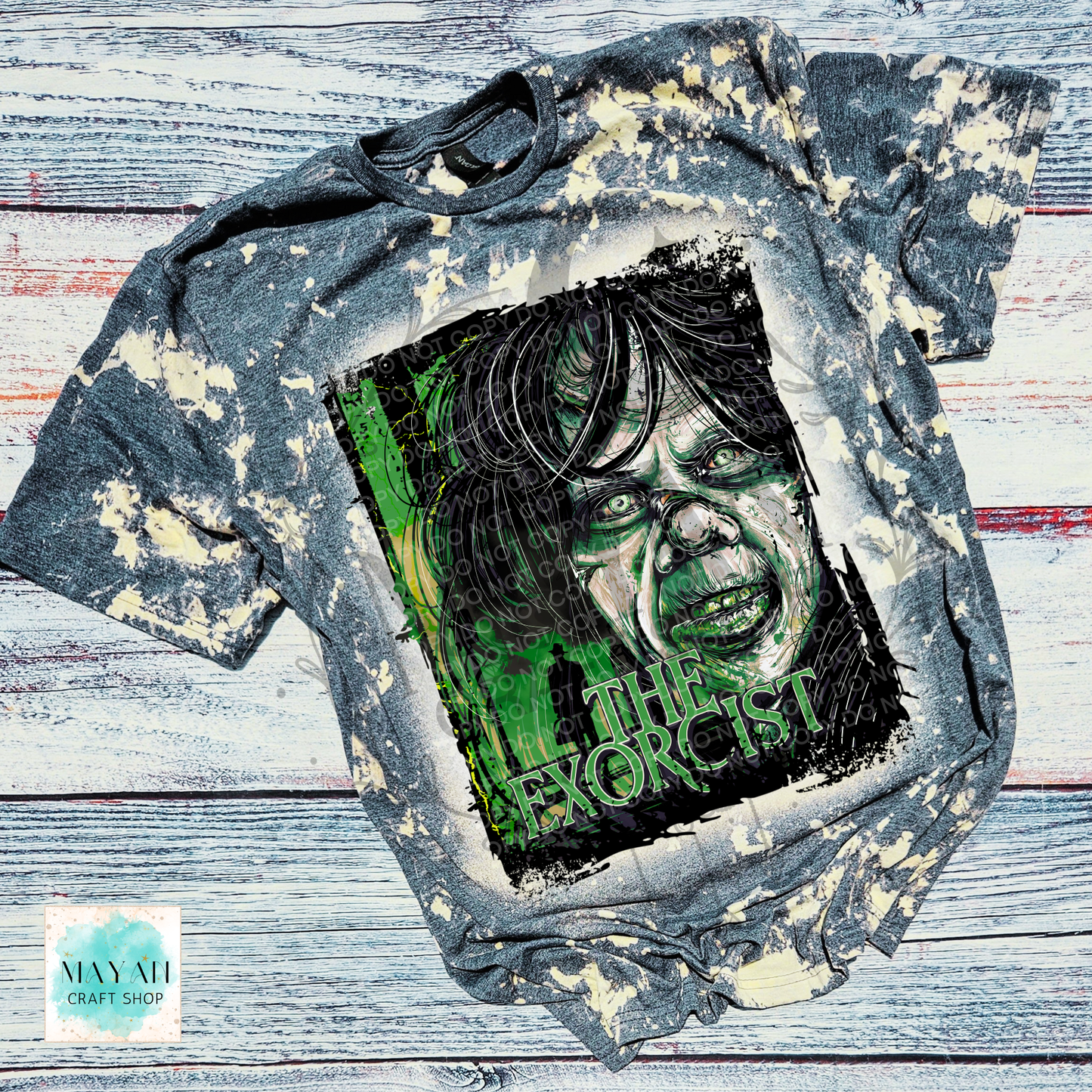 A short-sleeve graphic T-shirt laid flat on a distressed whitewashed wood background. The shirt is charcoal gray with a bleach-splatter finish and features a large rectangular front graphic showing a screaming face in black and green with a text.