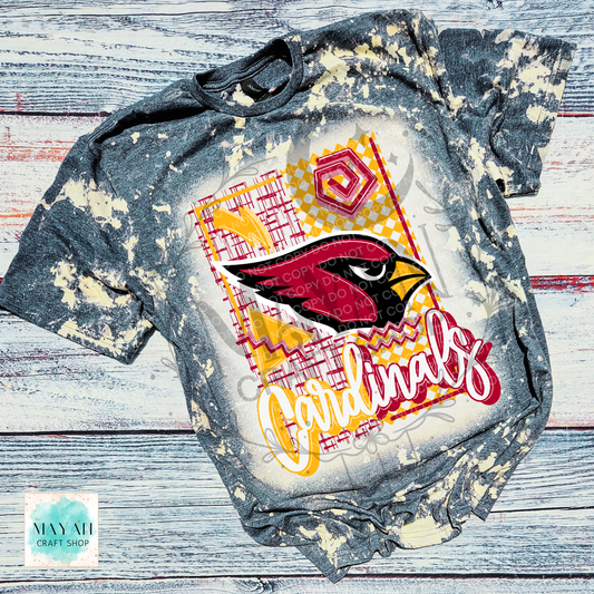 A short-sleeve graphic T-shirt laid flat on a distressed white and gray wood background. The shirt a heather charcoal bleached with a large front graphic featuring a football team logo, red and yellow geometric patterns.