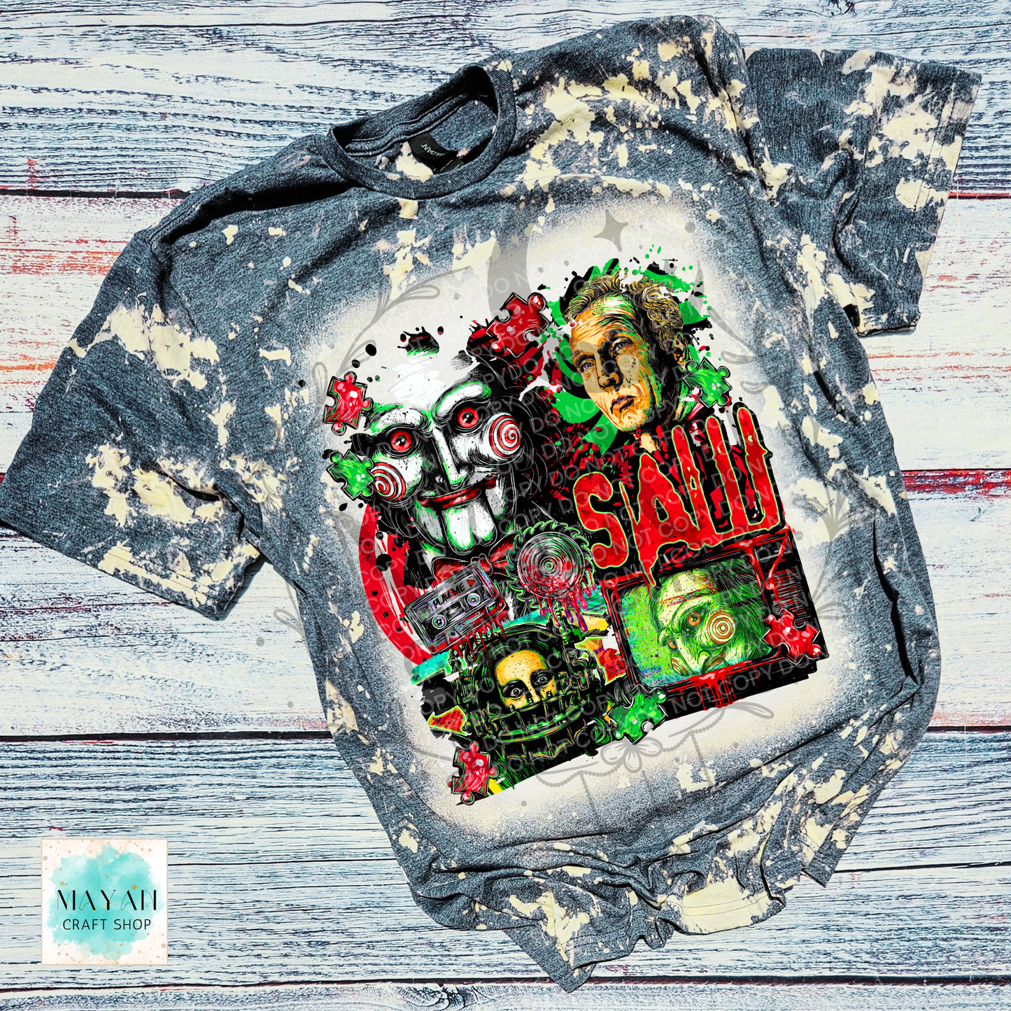 A short-sleeve graphic T-shirt laid flat on a rustic whitewashed wood surface. The shirt has a distressed bleach-splatter finish in gray and off-white, and the front features a large multicolor horror-themed print with a word in red and green, illustrated faces, skulls, and red roses.