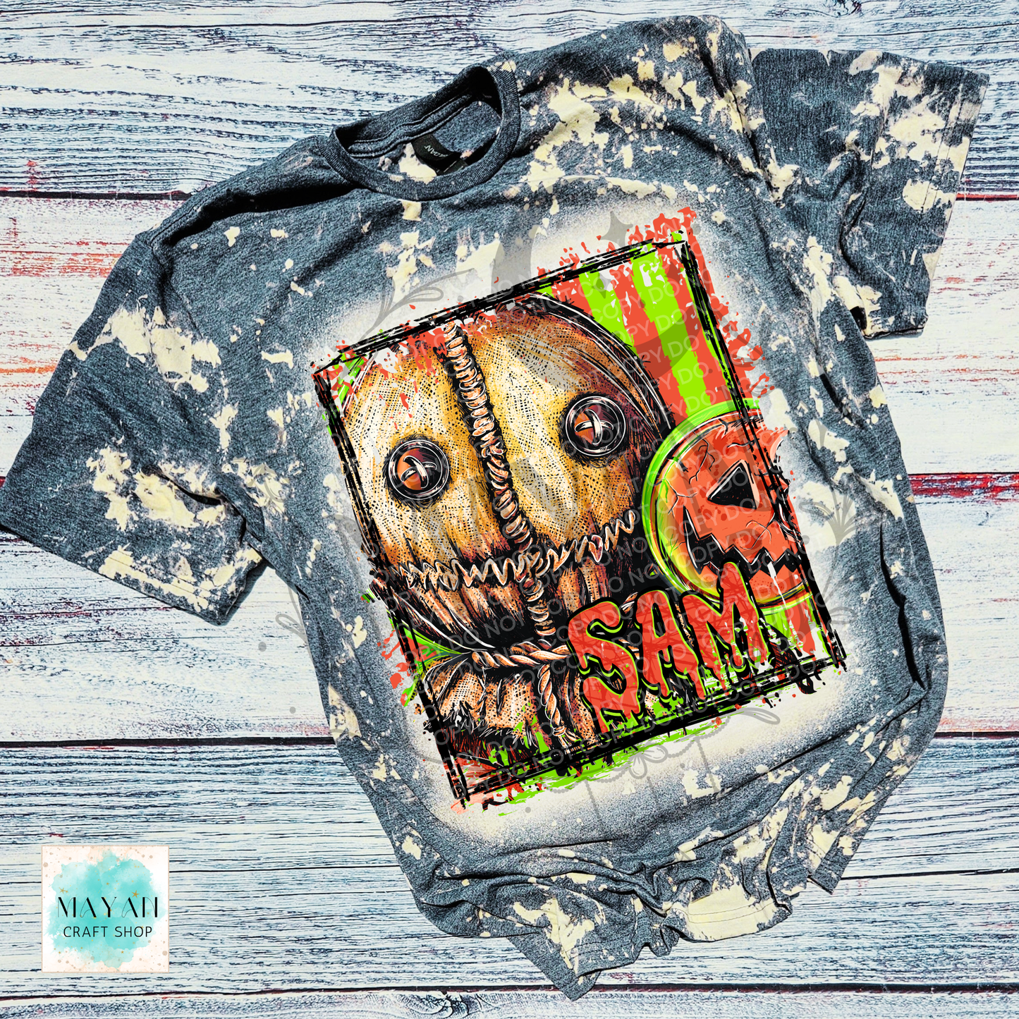 A short-sleeve graphic T-shirt laid flat on a distressed whitewashed wood background. The shirt is charcoal gray with a bleach-splatter finish and features a large front graphic of a stitched scarecrow face with a word in red.