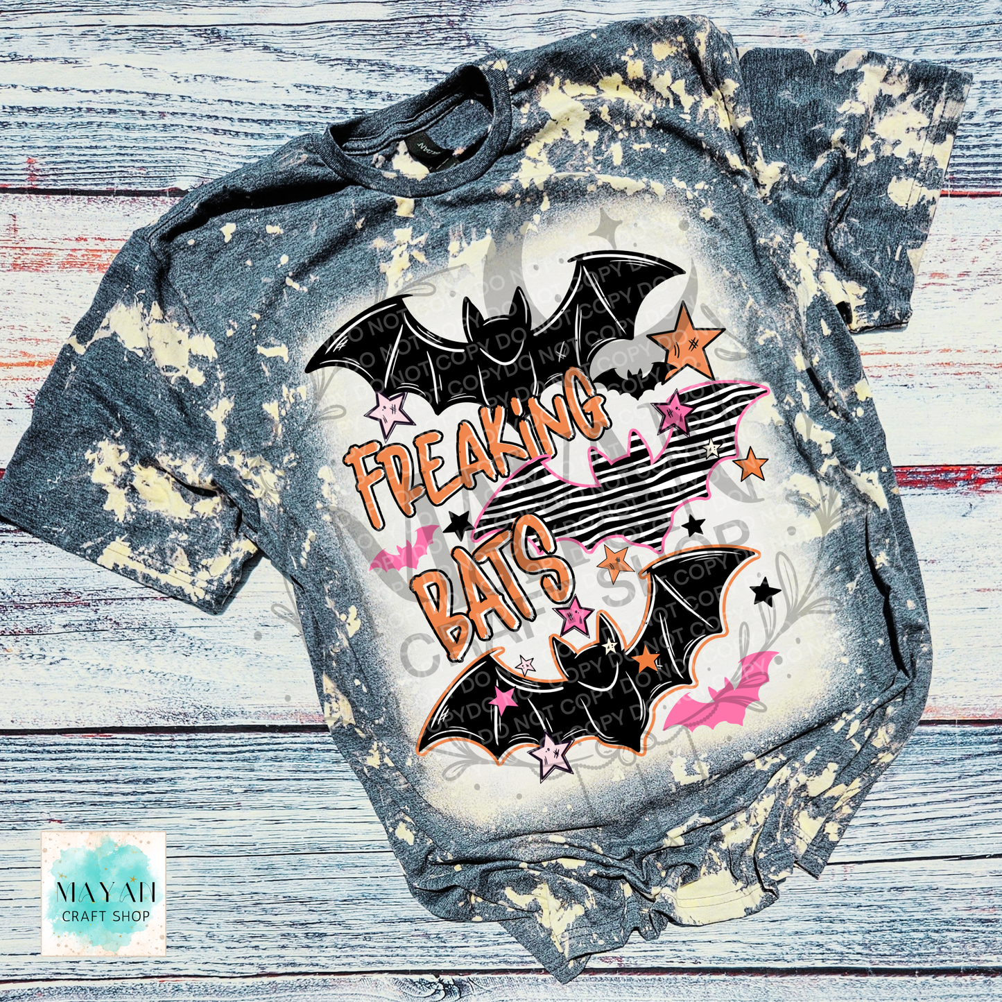 A short-sleeve graphic T-shirt laid flat on a distressed whitewashed wood background. The shirt has a dark gray bleach-splatter finish with a large front graphic featuring two black bats, pink and orange stars, and the words “FREAKING BATS” in orange.