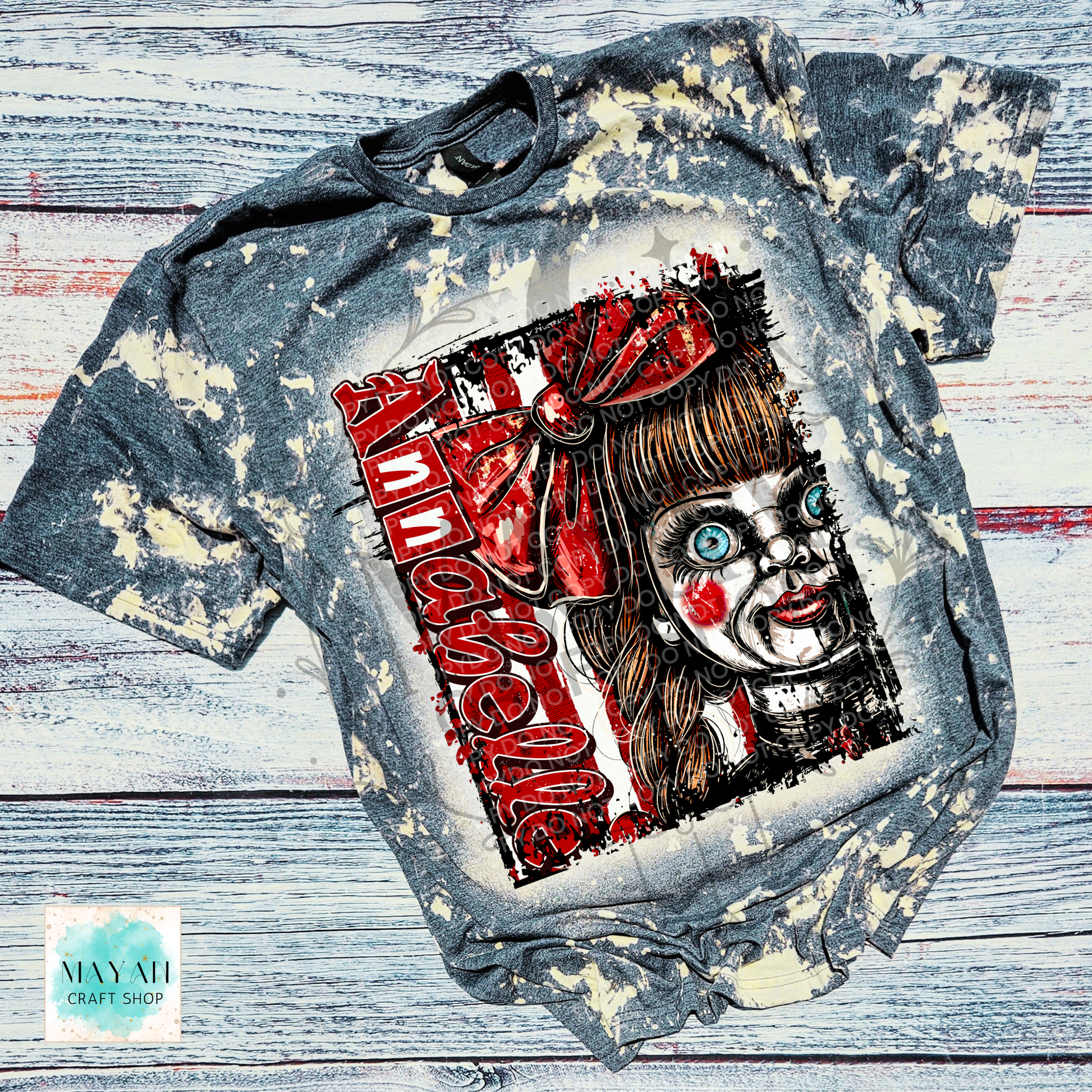 A short-sleeve graphic t-shirt with a bleached, distressed gray and white finish, featuring a large graphic print of a creepy doll character with a text. The shirt is laid flat on a rustic, whitewashed wooden plank background.