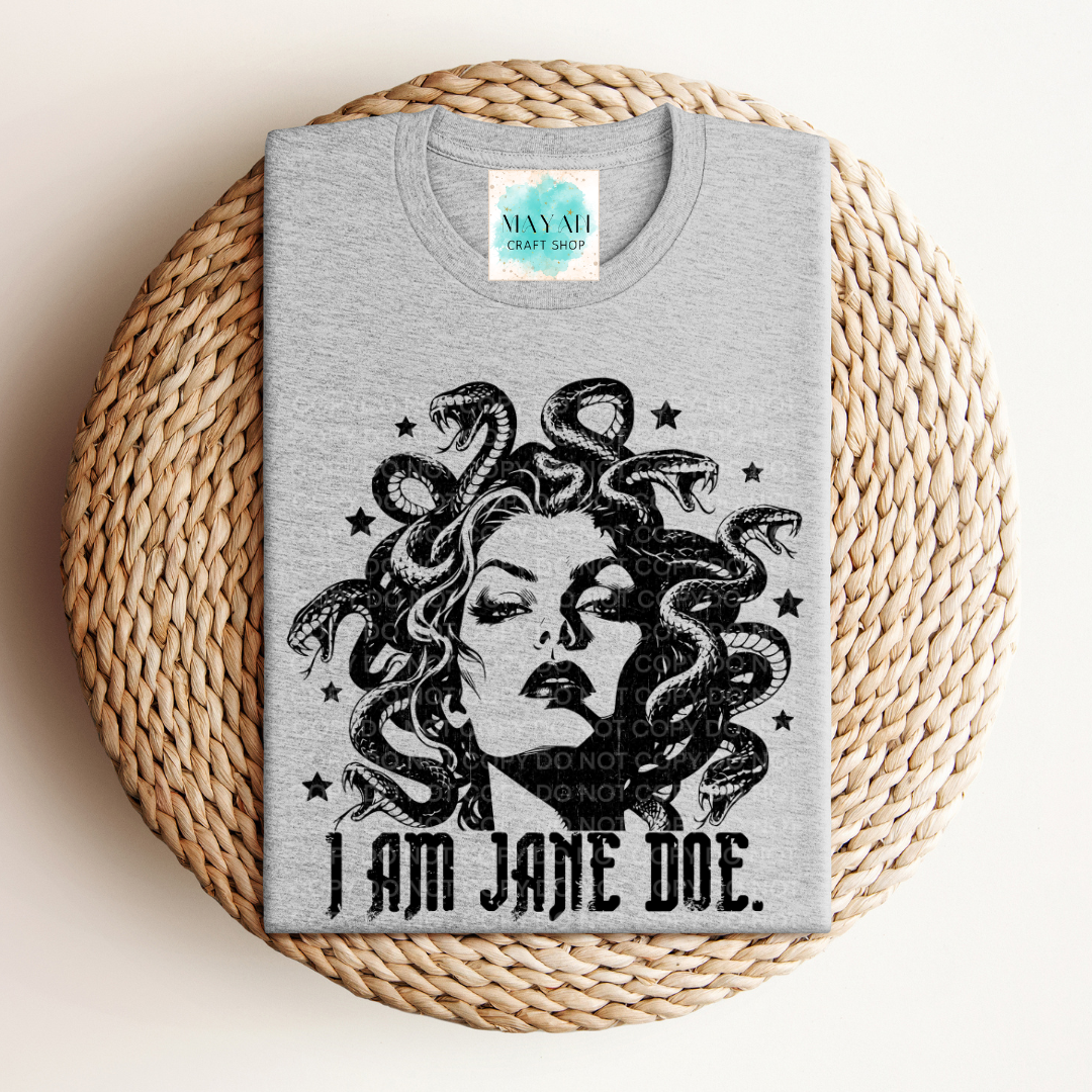 A folded heather gray t-shirt with a black graphic of Medusa and the text 'I AM JANE DOE.' is displayed on a round woven straw mat.