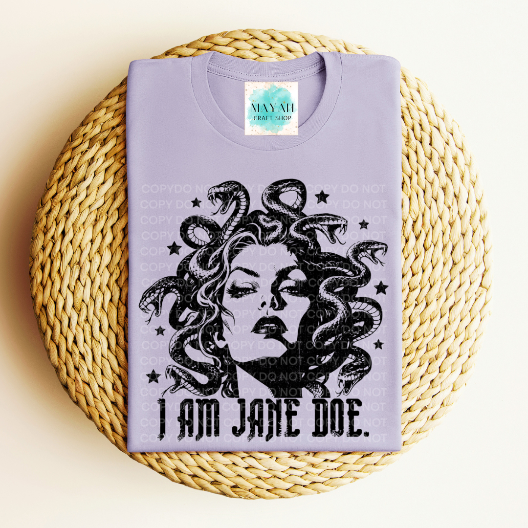 A lavender crewneck t-shirt featuring a black graphic of Medusa with the text 'I AM JANE DOE.' printed below it, displayed flat on a round woven straw mat.