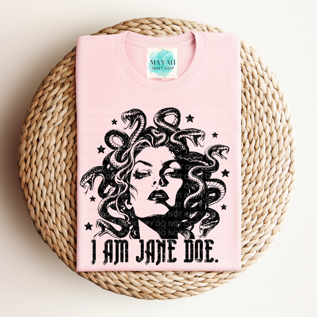A folded light pink T-shirt with a black graphic of Medusa and the text 'I AM JANE DOE.' is displayed on a round woven straw mat against a light beige background.