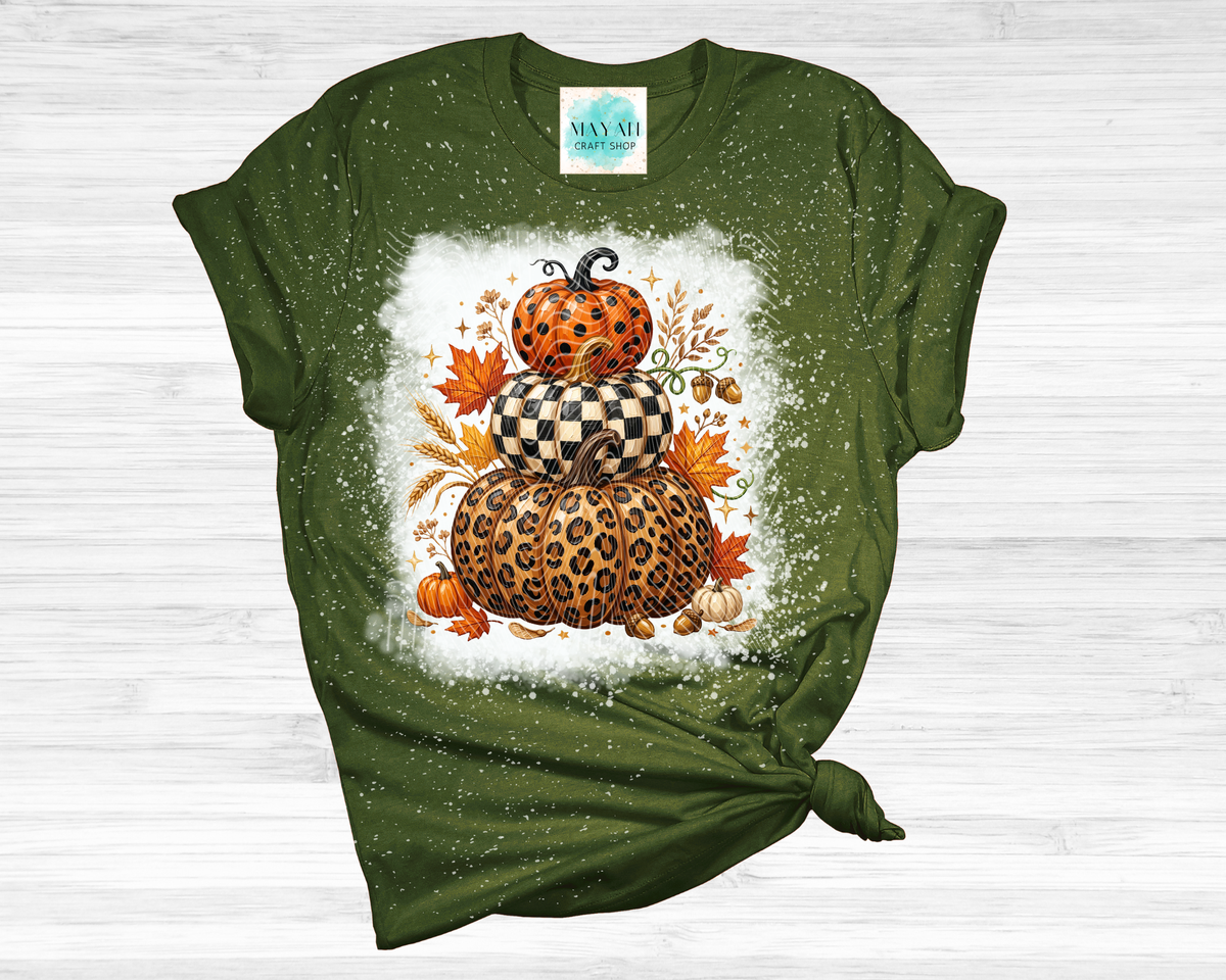 A heather forest green short-sleeve t-shirt with a knotted hem, featuring a central autumn graphic of stacked pumpkins in leopard, checkered, and orange patterns, surrounded by maple leaves and pinecones on a white splattered background. 