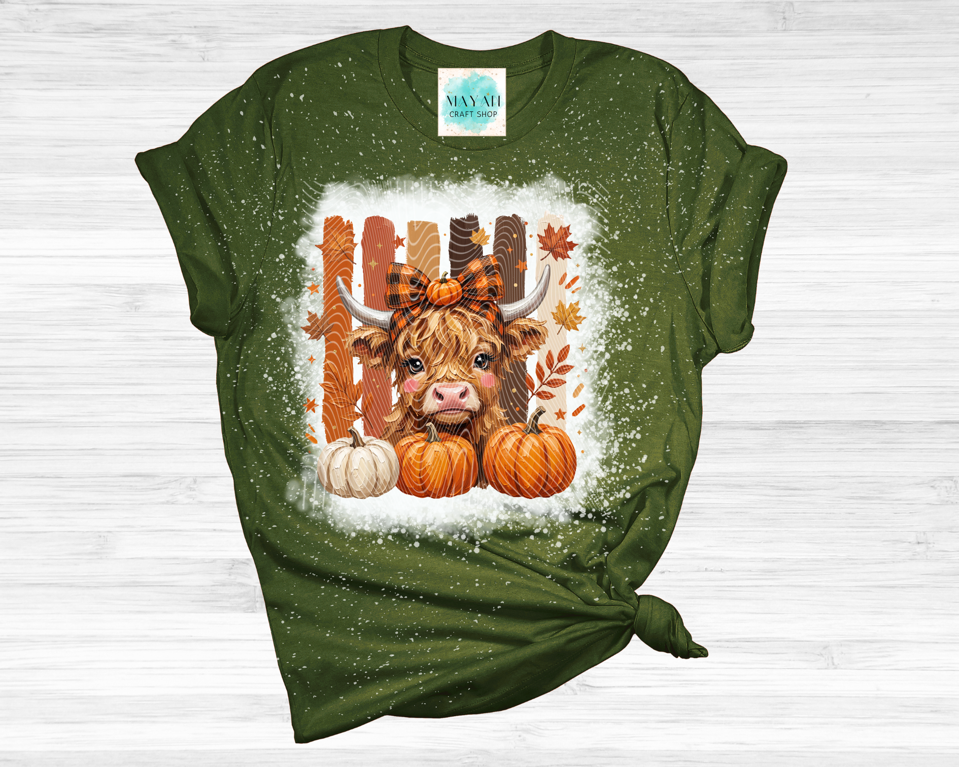 A heather forest green short-sleeve t-shirt with a distressed texture, featuring a central graphic of a Scottish Highland cow surrounded by pumpkins, autumn leaves, and a bow tie, set against a white splattered background. The shirt is styled with a knot at the bottom right and is laid flat on a light gray wood background.