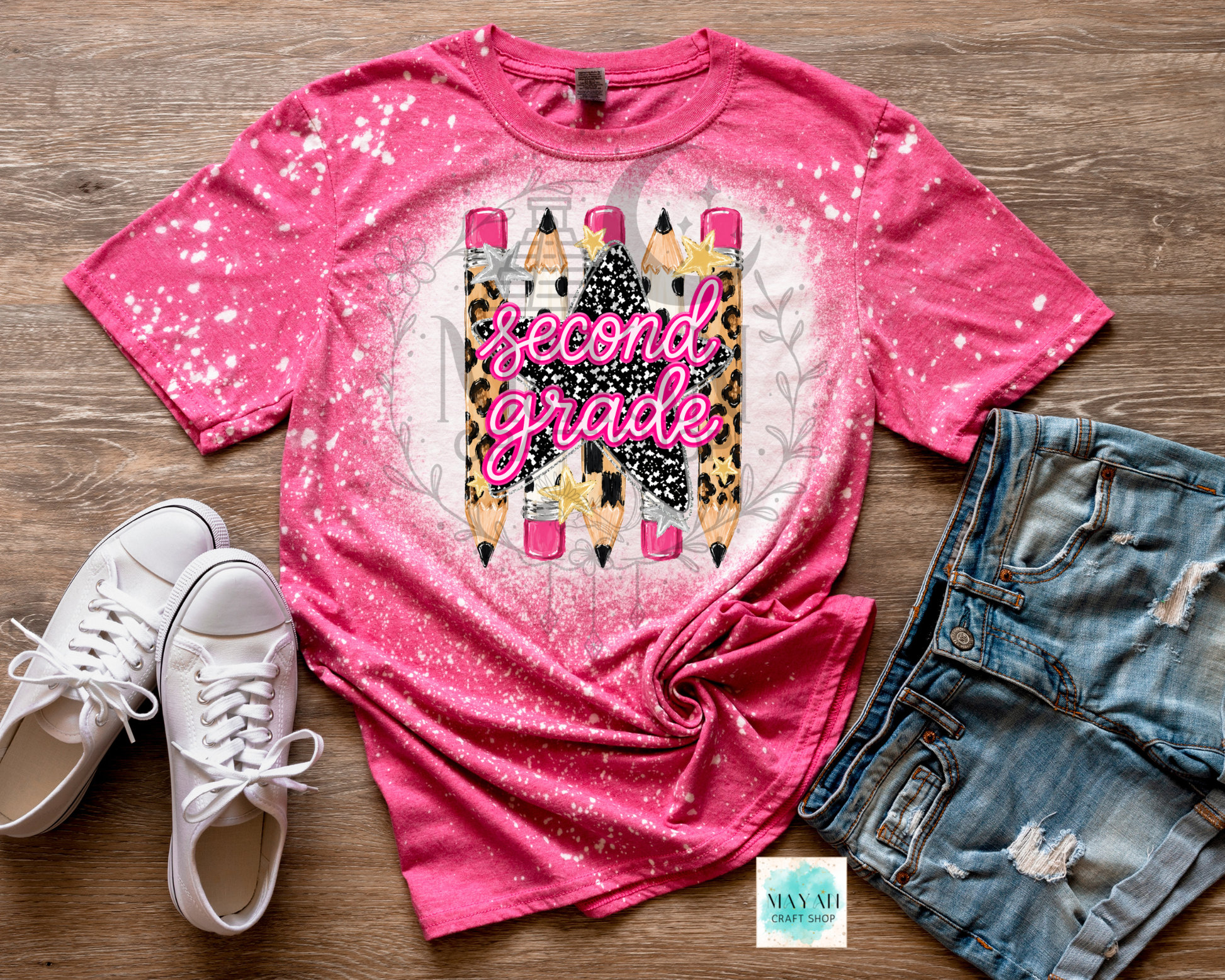 A pink short-sleeve graphic T-shirt laid flat on a wood surface, styled with white sneakers and distressed denim shorts. The shirt has a white splatter/gradient effect and a central graphic of colorful pencils with the words 'second grade' in pink script.