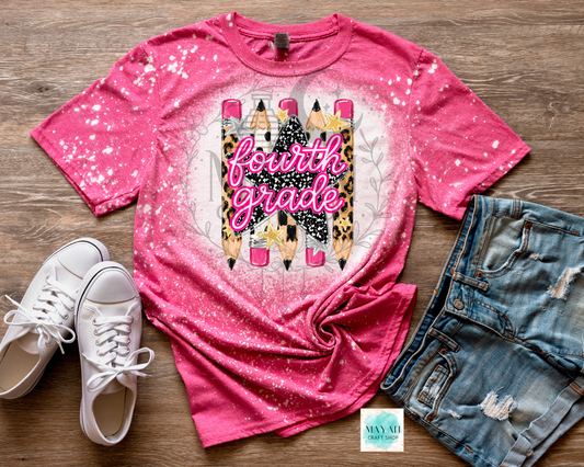 A pink short-sleeve graphic T-shirt laid flat on a wood surface, styled with white sneakers and distressed denim shorts. The shirt has a glittery pink and white gradient, a pencil illustration with the words 'fourth grade' in script.