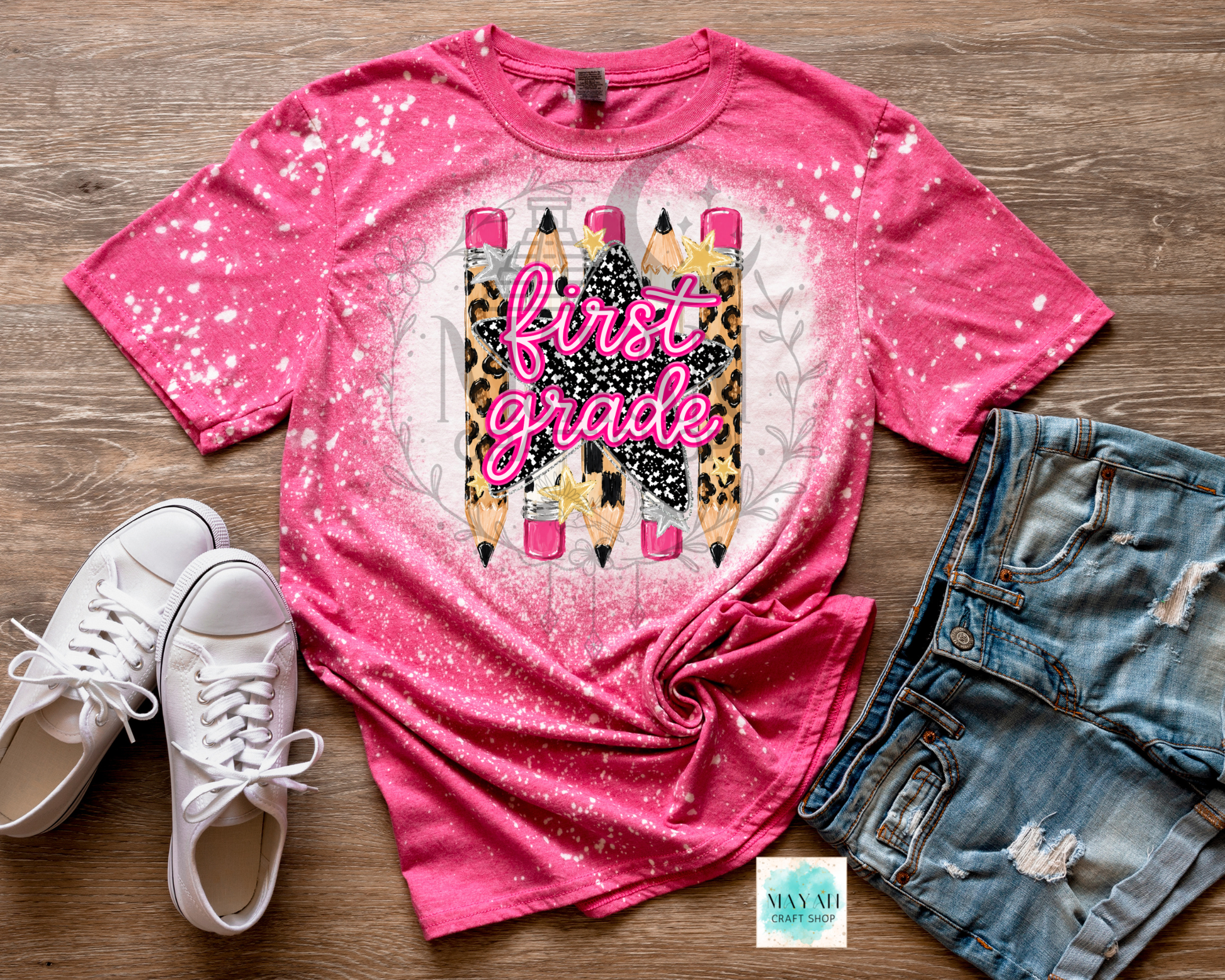 A pink short-sleeve graphic T-shirt laid flat on a wood surface, styled with white sneakers and distressed denim shorts. The shirt has a glittery pink-to-white gradient and a central graphic featuring three pencils in leopard print, black-and-white pattern, and pink, with the words 'first grade' in script.