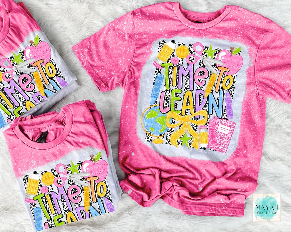Three pink short-sleeve graphic T-shirts arranged on a white faux-fur surface. The shirts feature a colorful retro-style 'Time To Learn' design with apples, pencils, and a notebook.