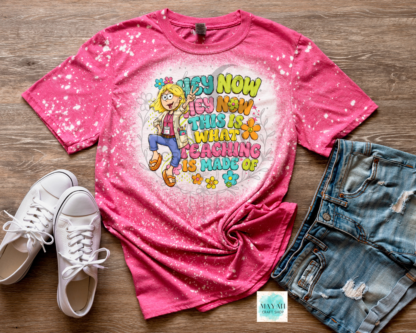 A pink short-sleeve graphic T-shirt laid flat on a wood surface, styled with white sneakers and distressed denim shorts. The shirt has a white splatter effect and a colorful retro-style graphic with the text 'HEY NOW I'M NOW I'M NOT TEACHING AS A MADE OF' and a cartoon girl.