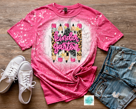 A pink short-sleeve graphic T-shirt laid flat on a wood surface, styled with white sneakers and distressed denim shorts. The shirt has a splatter/glitter finish and a large front graphic showing pencils in leopard print and black-and-white pattern with the words kinder garden' in pink script.