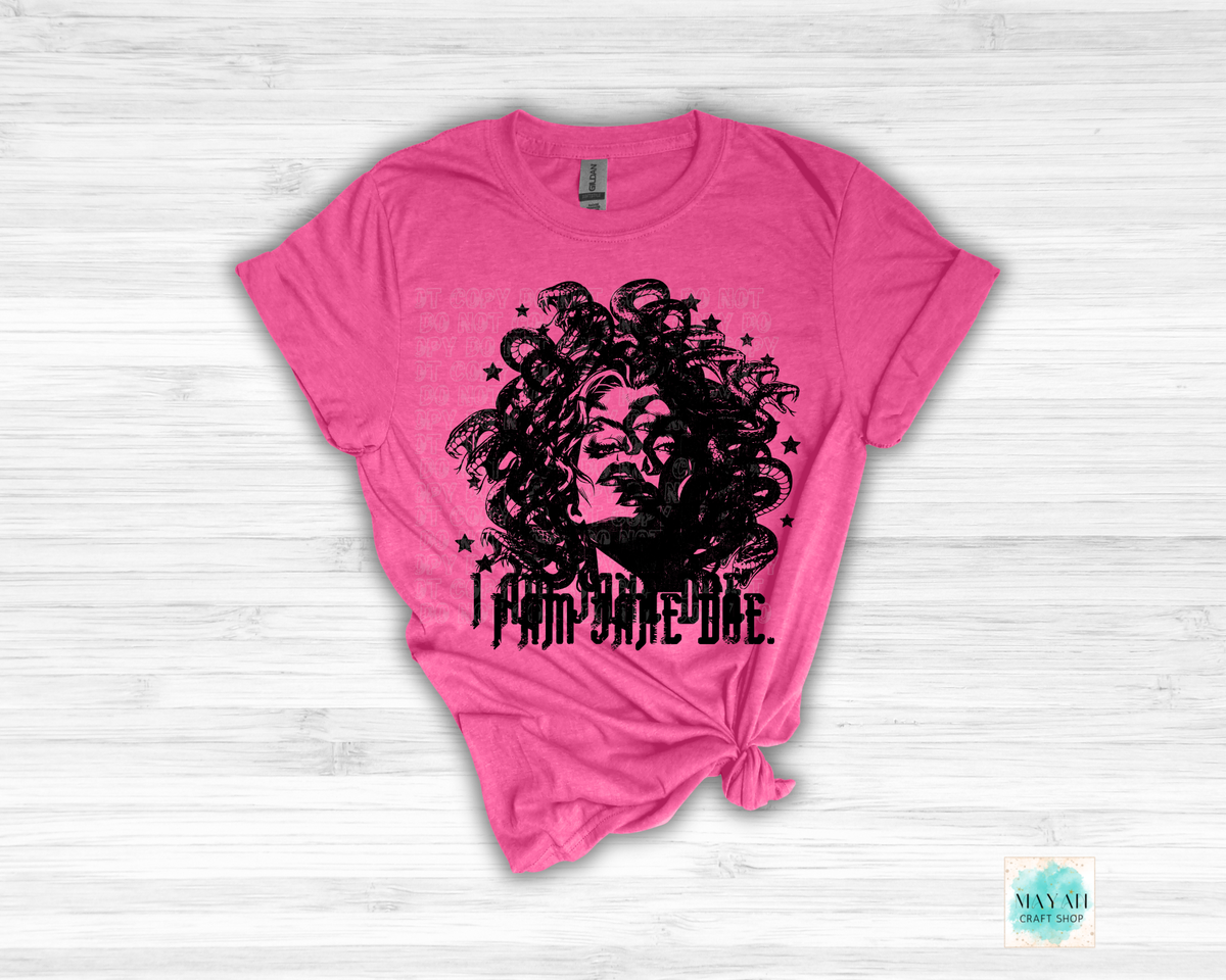 A heather heliconia short-sleeve t-shirt with a black graphic of a woman's face surrounded by stars and the text 'I AM Jane Doe.' printed on the front, styled with a knot at the bottom hem against a white wood plank background.